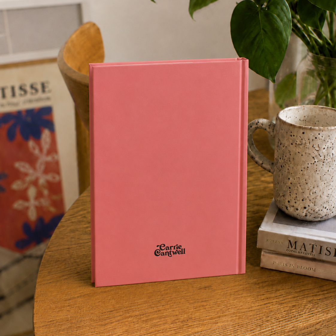 A pink hardcover book or notebook standing upright on a wooden table, with the title 'Carrie Cantwell' printed near the bottom. The scene is styled with a speckled ceramic mug on stacked books, a green plant, and framed art in the background.
